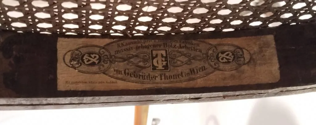 Close-up of an early Thonet paper label on the underside of a cane seat chair, reading Gebrüder Thonet Wien, with ornate lettering and decorative borders.