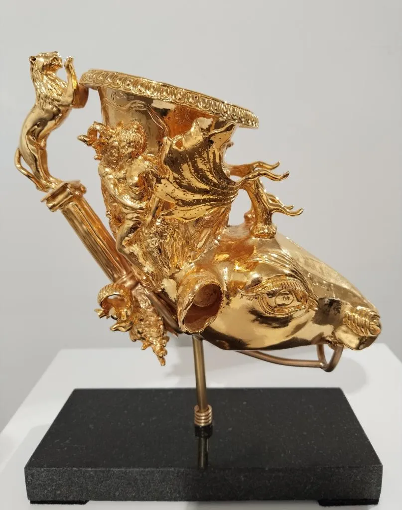 Gold ceremonial drinking vessel shaped as a ram’s head, with elaborate mythological reliefs and a lion-shaped handle, mounted on a black base.