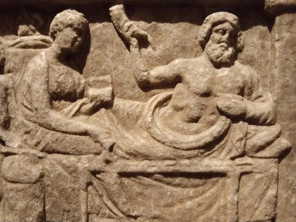 Marble relief showing a reclining male figure at a banquet, holding a rhyton with missing protome, accompanied by a seated female figure; Greek, 4th century BC.