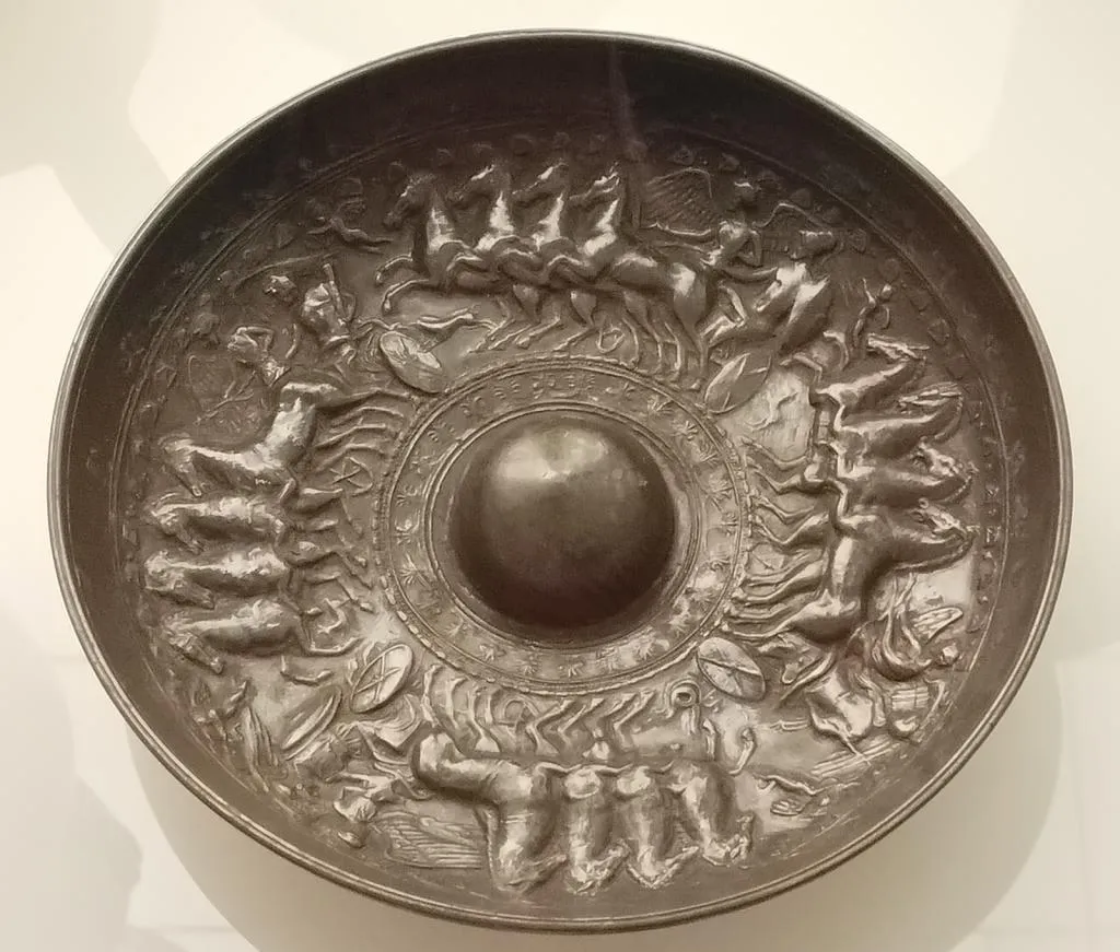 A round ancient ceramic phiale with a central omphalos and detailed relief decoration depicting Herakles ascending to Olympus in a chariot, surrounded by mythological figures.