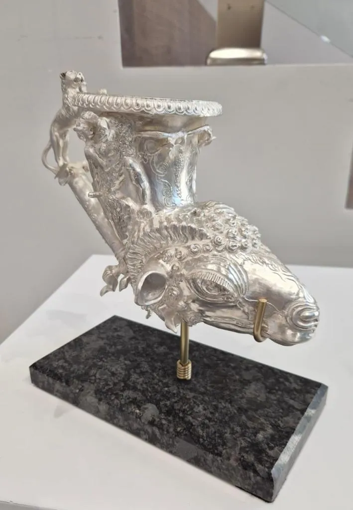 A silver-colored ritual drinking vessel with a ram head base and a lion-shaped handle, featuring seated mythological figures in relief on the neck.
