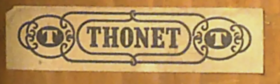 Close-up of a simplified Thonet paper label on wood, showing THONET in bold black letters flanked by decorative T emblems.