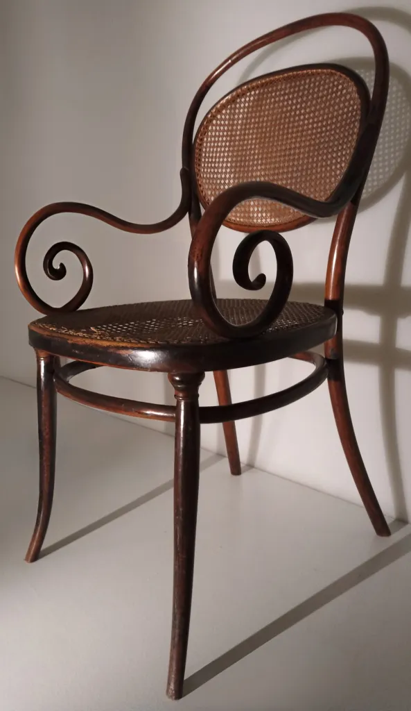 Seitenansicht des Thonet-Bugholzsessels Modell Nr. 11 mit ovaler Rückenlehne aus Rohrgeflecht, gebogenem Stützring unter dem Sitz und auffälligen, spiralförmigen Armlehnen.