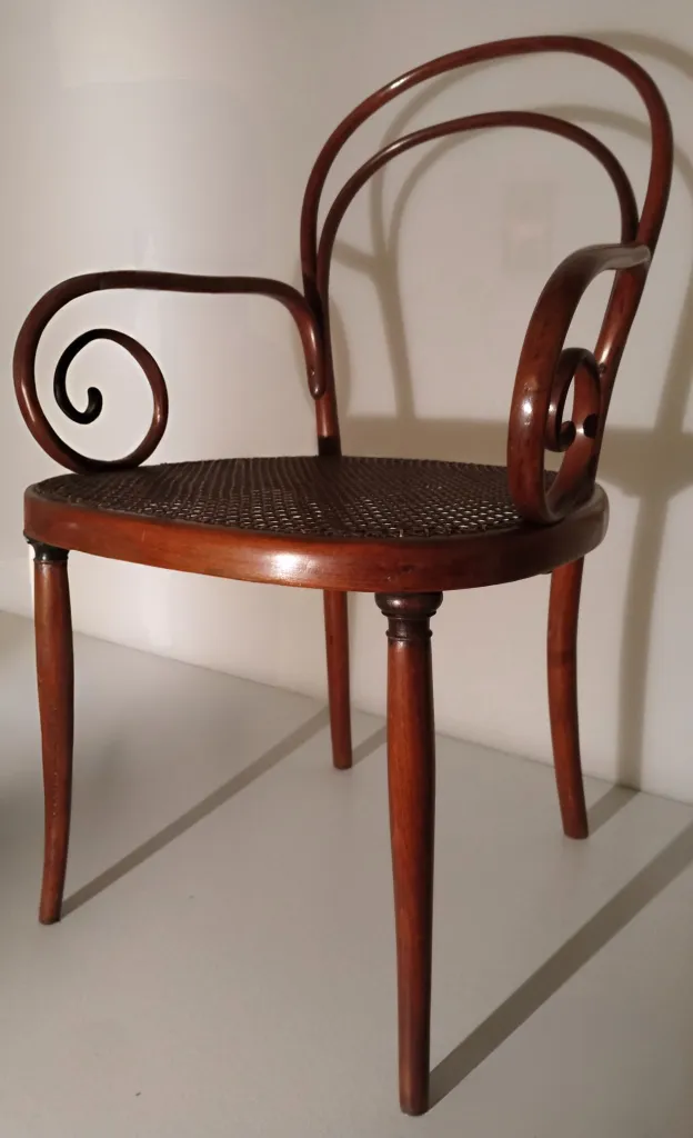 Seitenansicht des Thonet-Bugholzsessels Modell Nr. 8 mit rundem Rohrgeflechtsitz, gedrechselten Vorderbeinen und spiralförmigen Armlehnen, die in die doppelt gebogene Rückenlehne übergehen.