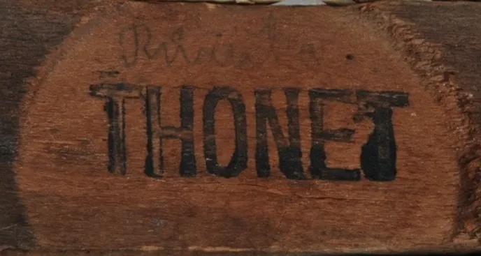 Close-up of a flat black ink stamp reading THONET on a wooden seat frame, showing wear and uneven ink application.