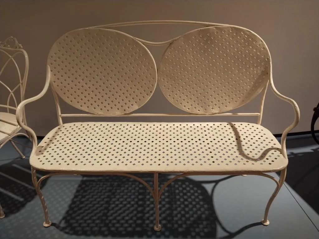 Photograph of a Thonet metal garden settee with two oval back panels, perforated seat, and curved arms, later repainted in white.