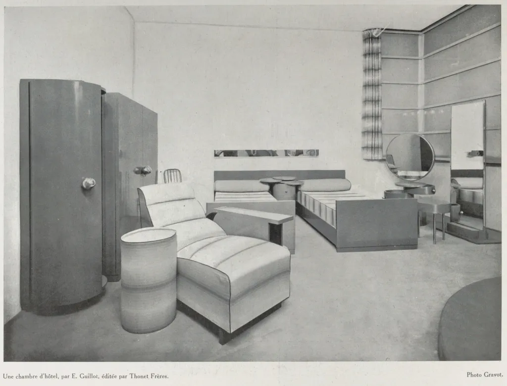 Black-and-white photograph of a 1929 hotel room designed by E. Guillot and furnished by Thonet Frères, featuring a lounge chair, bed, wardrobe, and other modern furniture.